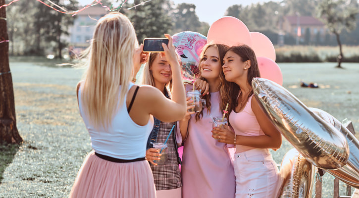 Grupa młodych kobiet celebruje 18 urodziny nad wodą z balonami i drinkami, robiąc sobie zdjęcia.