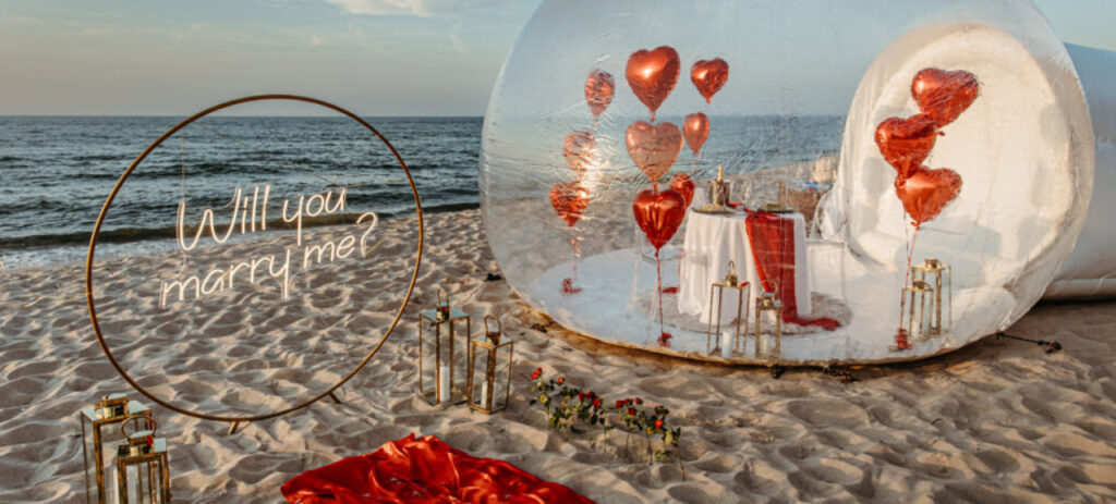Sceneria zaręczyn na plaży nad morzem - napis 'Will you marry me?' na piasku, czrwone balony w kształcie serc, płatki róż rozrzucone na piasku i romantyczna dekoracja na tle zachodzącego słońca.