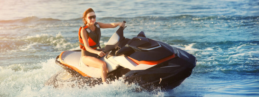 Uśmiechnięta kobieta w kamizelce ratunkowej pędząca skuterem wodnym po jeziorze.