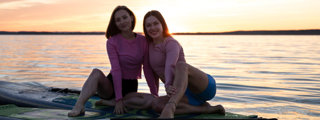 Przyjaciółki w różowych bluzach odpoczywające na SUP nad spokojnym jeziorem podczas zachodu słońca.