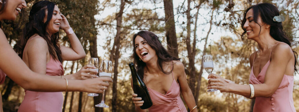 Grupa przyjaciółek w różowych sukienkach świętująca wieczór panieński z butelką szampana w plenerze.